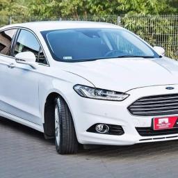 Wypożyczalnia samochodów MakaraCars - Biały samochód Ford Mondeo na tle zieleni w Bełchatowie, z widoczną tabliczką reklamową wypożyczalni na przednim zderzaku.