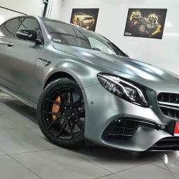 Wypożyczalnia samochodów MakaraCars - Matowy, srebrny Mercedes-Benz E63 AMG z czarnymi felgami i pomarańczowymi zaciskami hamulcowymi, zaparkowany w dobrze oświetlonym salonie samochodowym z plakatami motoryzacyjnymi na ścianie.