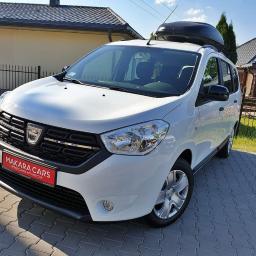 Wypożyczalnia samochodów MakaraCars - Biały samochód marki Dacia z bagażnikiem dachowym na tle domu jednorodzinnego, na czerwonej tabliczce widnieje napis MAKARA CARS.