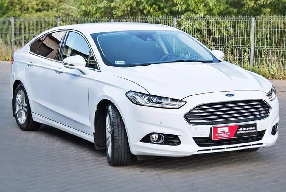 Biały samochód Ford Mondeo na tle zieleni w Bełchatowie, z widoczną tabliczką reklamową wypożyczalni na przednim zderzaku.
