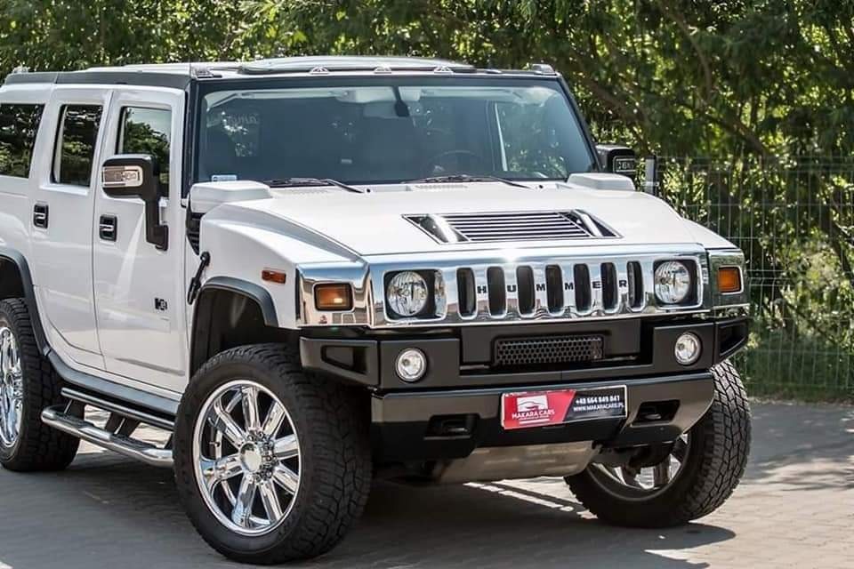 Biały Hummer H2 z chromowanymi felgami, prezentowany na zewnątrz z zielonymi drzewami w tle, widoczna tabliczka z nazwą firmy.
