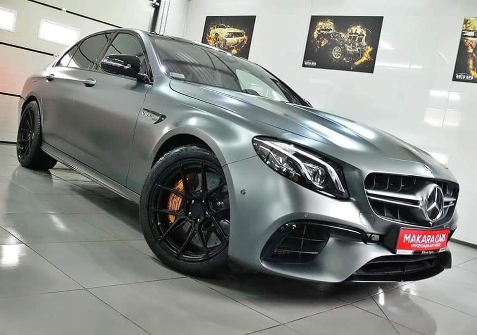 Matowy, srebrny Mercedes-Benz E63 AMG z czarnymi felgami i pomarańczowymi zaciskami hamulcowymi, zaparkowany w dobrze oświetlonym salonie samochodowym z plakatami motoryzacyjnymi na ścianie.