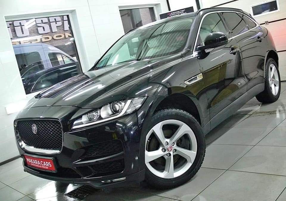 Błyszczący, czarny samochód Jaguar SUV, model F-Pace, zaparkowany w jasnym pomieszczeniu z białymi ścianami i kafelkami na podłodze; widoczny napis 'MAKARA CARS' na czerwonym tle na przednim zderzaku.