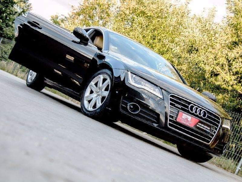 Czarny samochód osobowy marki Audi z otwartymi drzwiami kierowcy, widoczne aluminiowe felgi i fragment zieleni w tle.