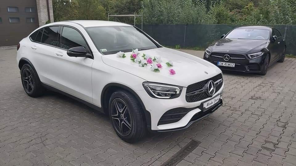Biały Mercedes GLC Coupe udekorowany różowymi i białymi kwiatami na masce, zaparkowany obok czarnego Mercedesa na brukowanym parkingu.