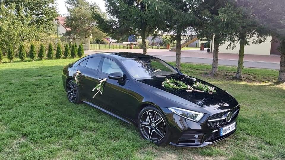 Czarny Mercedes-Benz CLS z dekoracjami kwiatowymi na masce i klamkach, zaparkowany na trawniku przed domem, prawdopodobnie przygotowany do ślubu.