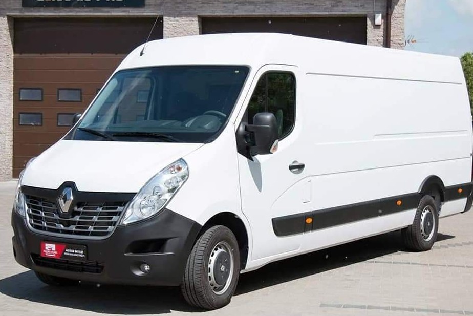 Biały samochód dostawczy Renault Master zaparkowany przed budynkiem z brązową bramą garażową.