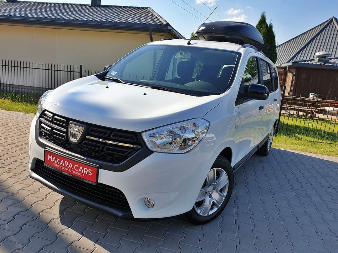 Biały samochód marki Dacia z bagażnikiem dachowym na tle domu jednorodzinnego, na czerwonej tabliczce widnieje napis MAKARA CARS.