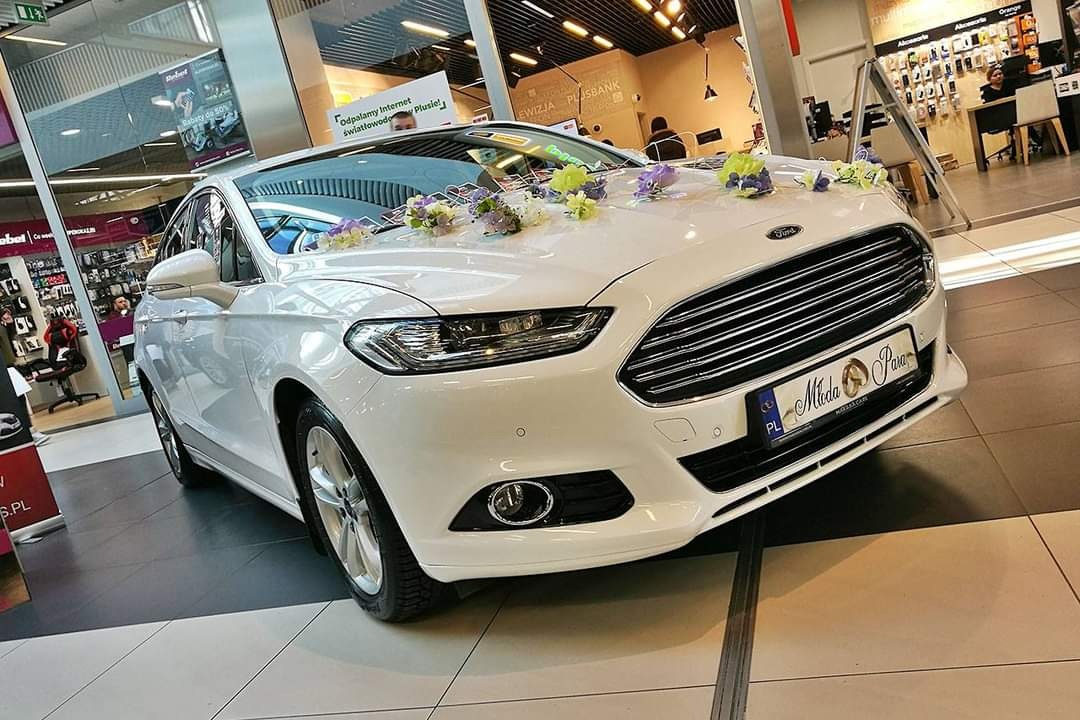 Biały Ford z dekoracjami kwiatowymi na masce, tablica rejestracyjna z napisem 'Młoda Para', zaparkowany wewnątrz centrum handlowego.