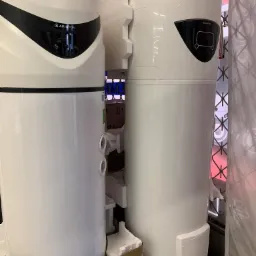 Dwa białe, fabrycznie zapakowane, pionowe podgrzewacze wody marki Ariston, widoczne elementy sterowania i obudowy, zabezpieczone styropianem i folią.