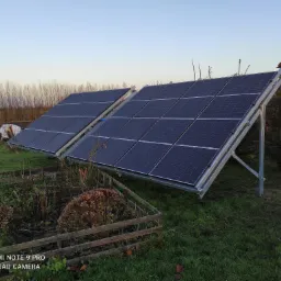 Dwa panele słoneczne zamontowane na metalowej konstrukcji na trawiastym terenie, z widocznym ogrodem warzywnym w tle i fragmentem domu z prawej strony.