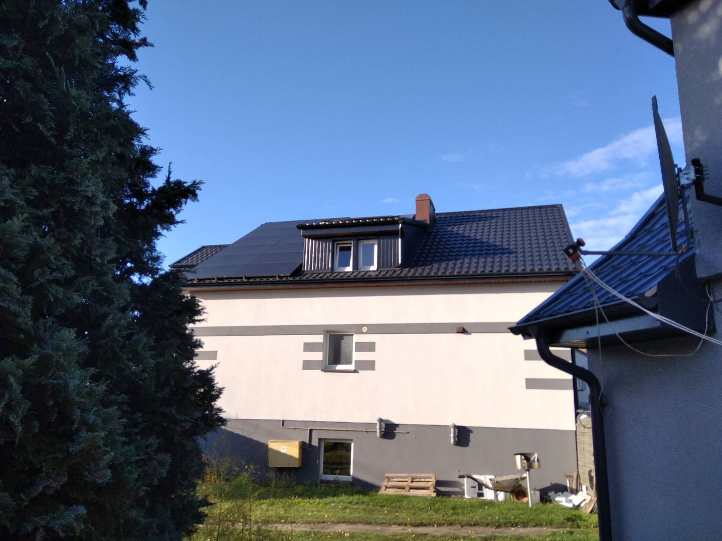 Dom jednorodzinny z panelami słonecznymi na dachu, widok z poziomu gruntu, częściowo zasłonięty przez drzewo iglaste po lewej stronie.