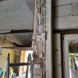 Surowe wnętrze z instalacją elektryczną: przewody, puszki, włączniki i gniazdo siłowe zamontowane na ścianie z bloczków betonowych, drabina aluminiowa oparta o ścianę, widoczne kable wystające ze...