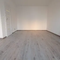 Pusty pokój po remoncie z panelami podłogowymi imitującymi drewno, białe ściany i białe drzwi z przeszkleniem, widoczne wystające kable ze ściany i sufitu.