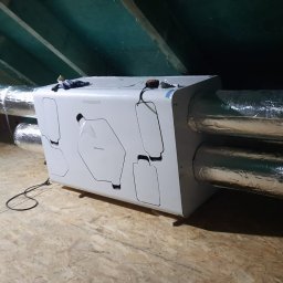 Mardom 13 Marcin Kozieł - Centrala wentylacyjna z rekuperacją zamontowana na poddaszu, widoczne przewody wentylacyjne w izolacji aluminiowej, narzędzia leżące na podłodze, surowe drewniane wykończenie poddasza.