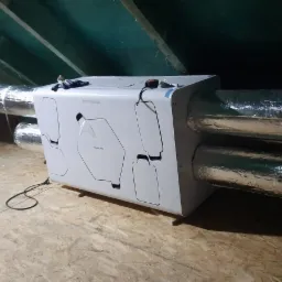 Centrala wentylacyjna z rekuperacją zamontowana na poddaszu, widoczne przewody wentylacyjne w izolacji aluminiowej, narzędzia leżące na podłodze, surowe drewniane wykończenie poddasza.