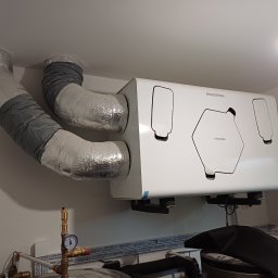 Mardom 13 Marcin Kozieł - Biała jednostka rekuperacyjna THESSLAGREEN zamontowana pod sufitem, z podłączonymi izolowanymi przewodami wentylacyjnymi, widoczne rury miedziane i manometr.