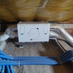 Mardom 13 Marcin Kozieł - Centrala wentylacyjna z rekuperacją, biała obudowa z czarnymi elementami, podwieszona pod stropem, widoczne niebieskie rury wentylacyjne i srebrne przewody izolowane termicznie, strop ocieplony...