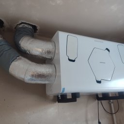 Mardom 13 Marcin Kozieł - Biała jednostka rekuperacyjna marki Thessla Green zamontowana pod sufitem, z podłączonymi aluminiowymi przewodami wentylacyjnymi w izolacji, widoczne niedokończone wykończenie ścian.