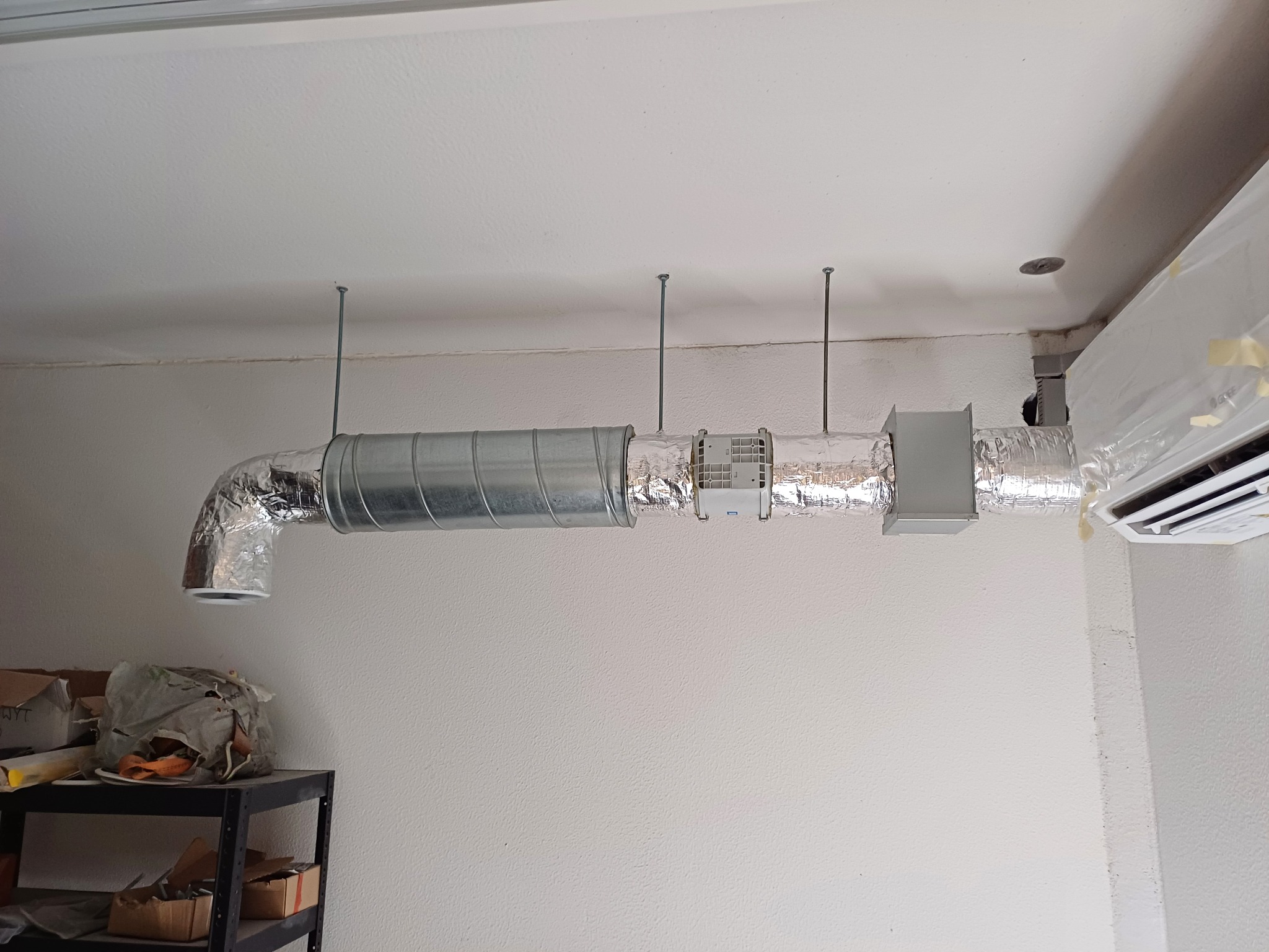 Instalacja wentylacji: widok na kanał wentylacyjny z metalu i aluminium, zawieszony pod sufitem na metalowych prętach, z klimatyzatorem w rogu pokoju, w tle widoczny regał z kartonami.