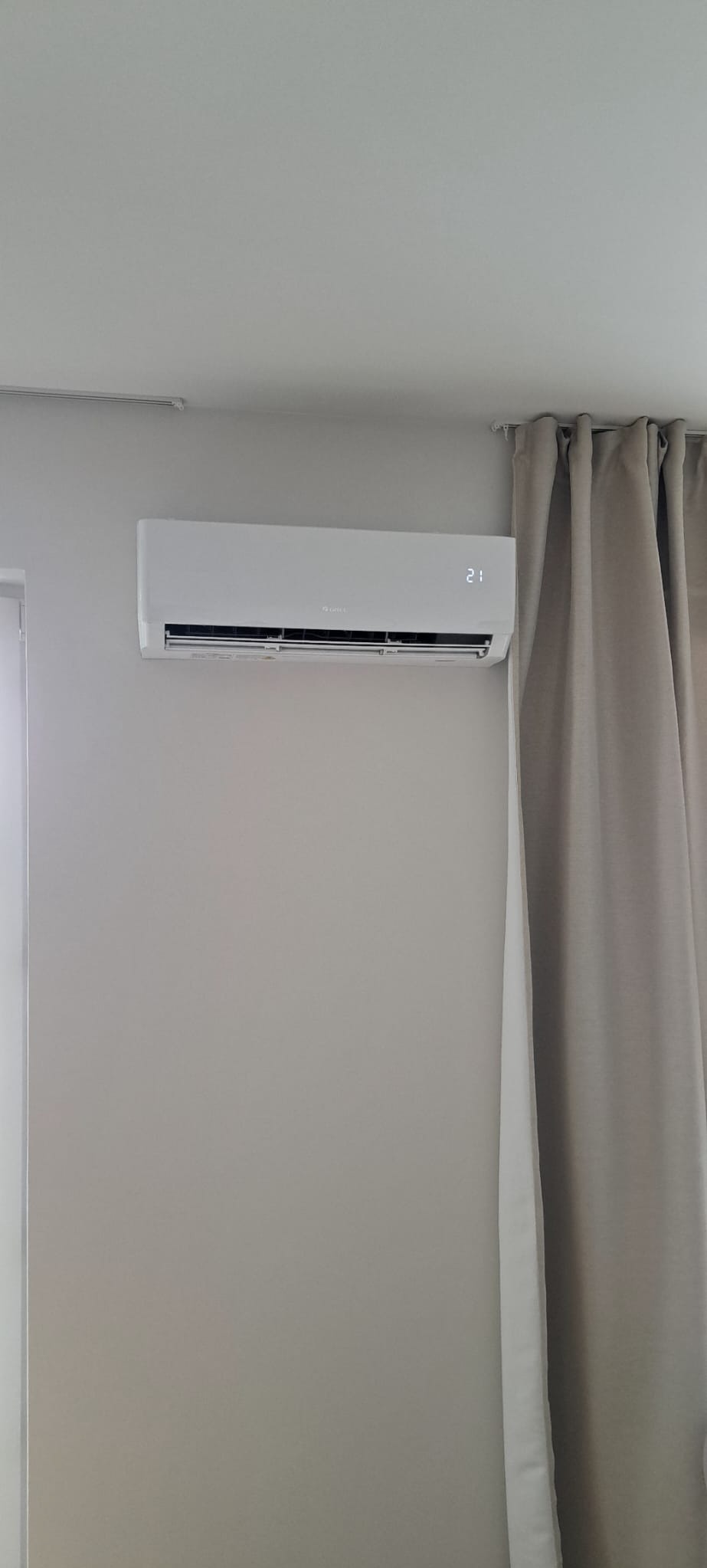 Biały klimatyzator ścienny z wyświetlaną temperaturą 21 stopni Celsjusza, zamontowany w jasnym pomieszczeniu obok zasłony w odcieniach szarości.