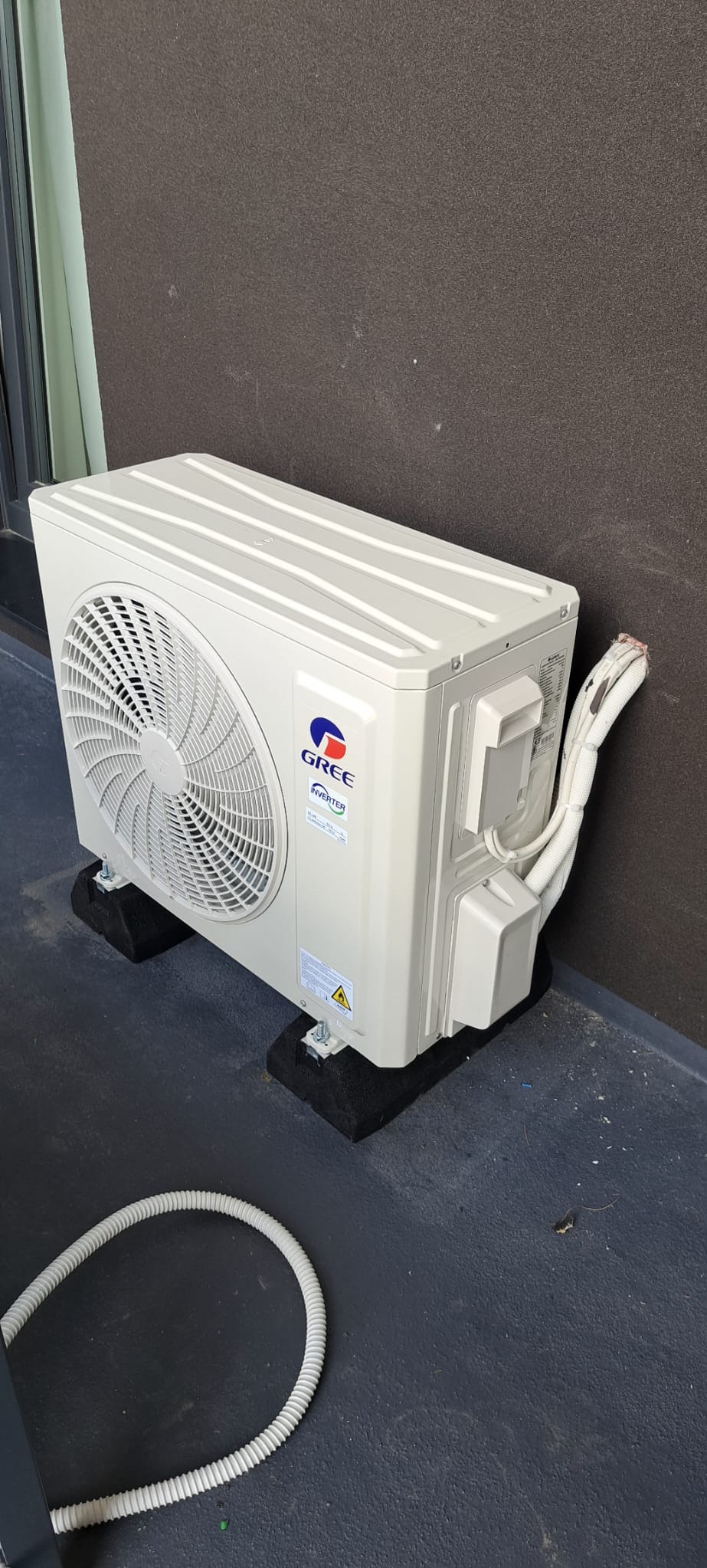 Zewnętrzna jednostka klimatyzacji marki Gree, zamontowana na czarnych podkładkach antywibracyjnych, z widocznymi przewodami i fragmentem białej rury odprowadzającej skropliny na szarym balkonie.