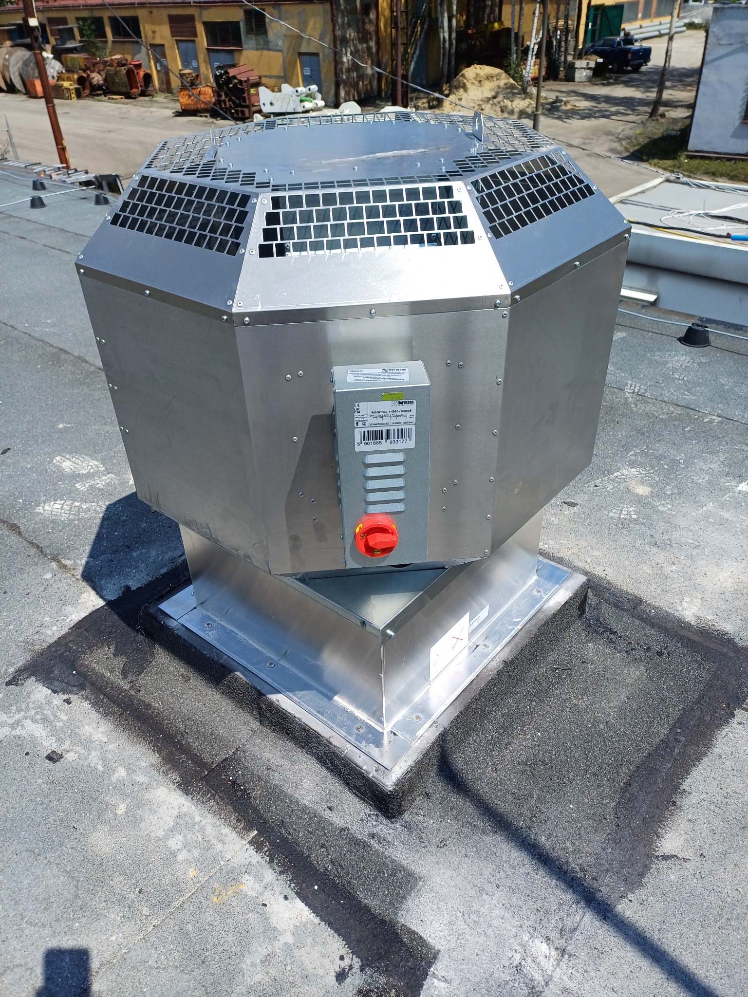 Srebrny, ośmiokątny wentylator dachowy z kratką ochronną, zamontowany na bitumicznej powierzchni dachu, widoczne elementy mocujące i etykieta producenta.