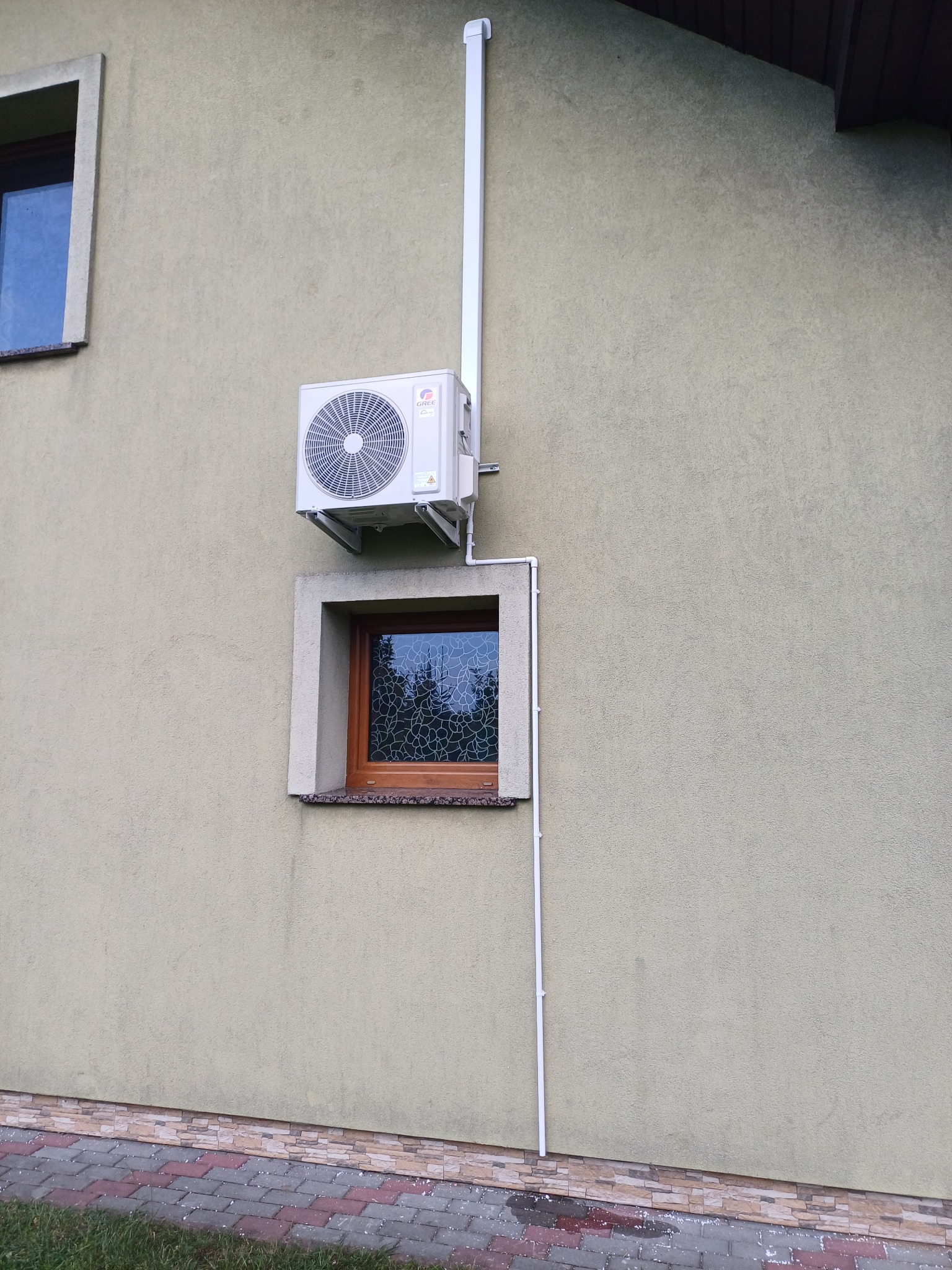 Zewnętrzna jednostka klimatyzacji Gree zamontowana na elewacji domu, rury odprowadzające skropliny prowadzone wzdłuż ściany obok okna z ozdobną szybą.