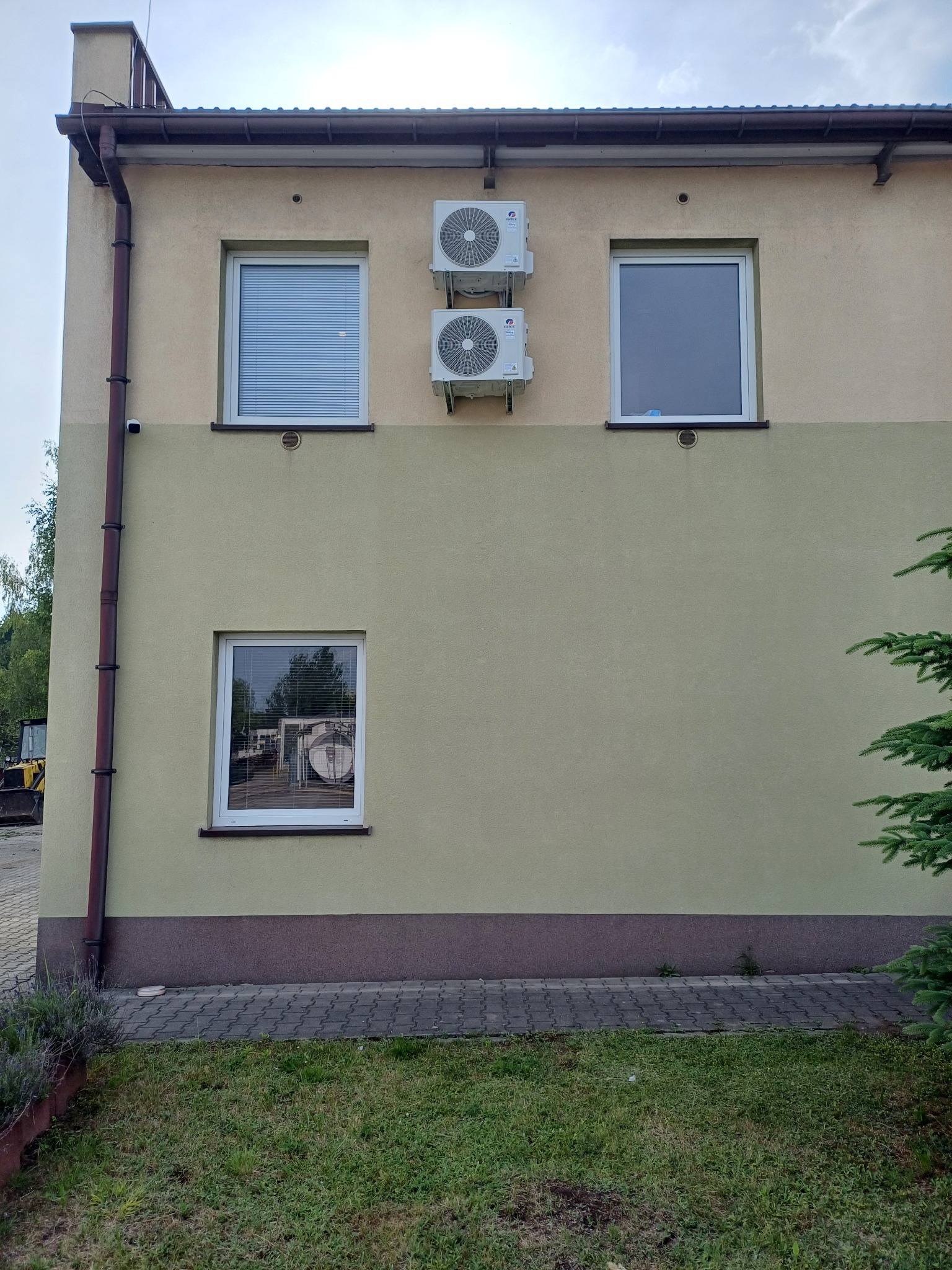Dwa agregaty klimatyzacyjne zamontowane na elewacji budynku o beżowo-oliwkowym kolorze, widoczne okna z roletami i bez rolet, rynna spustowa wzdłuż ściany.