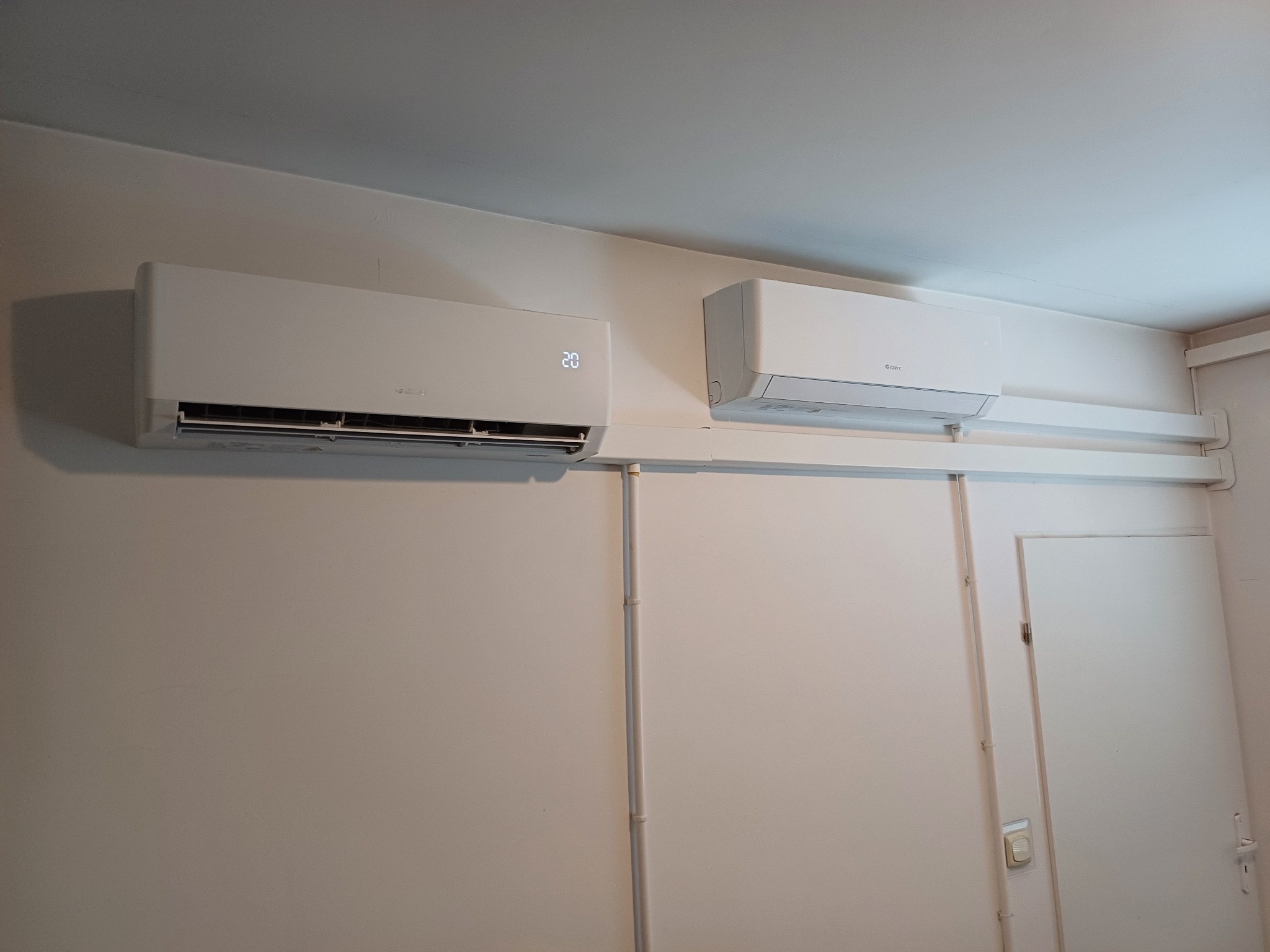 Dwa białe klimatyzatory marki Gree zamontowane na ścianie w pomieszczeniu. Jeden z wyświetlaczem pokazującym temperaturę 20 stopni Celsjusza.