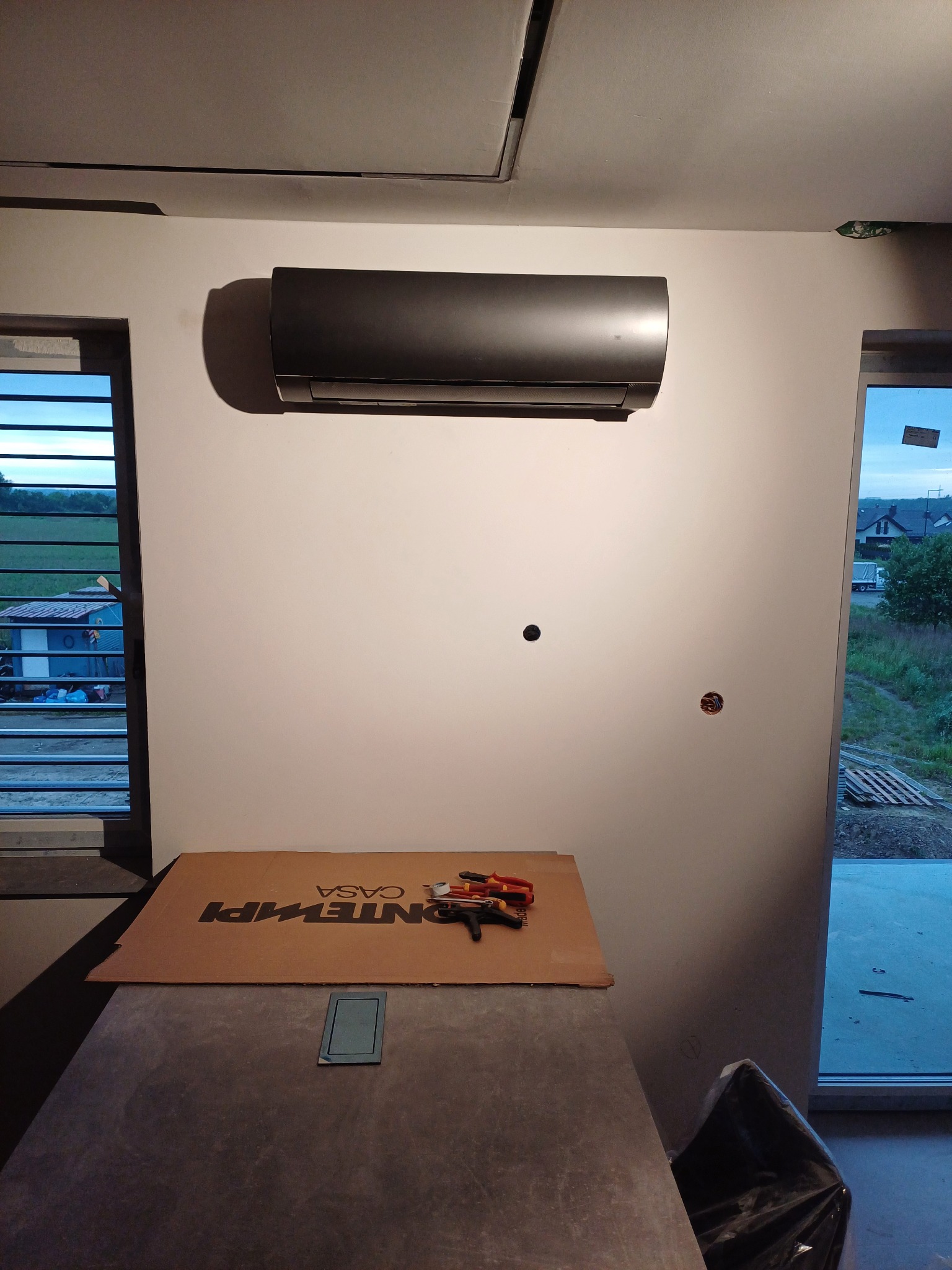 Czarny klimatyzator na białej ścianie w surowym wnętrzu, z narzędziami na kartonie, widoczne otwory na kable i okno z widokiem na zewnątrz.