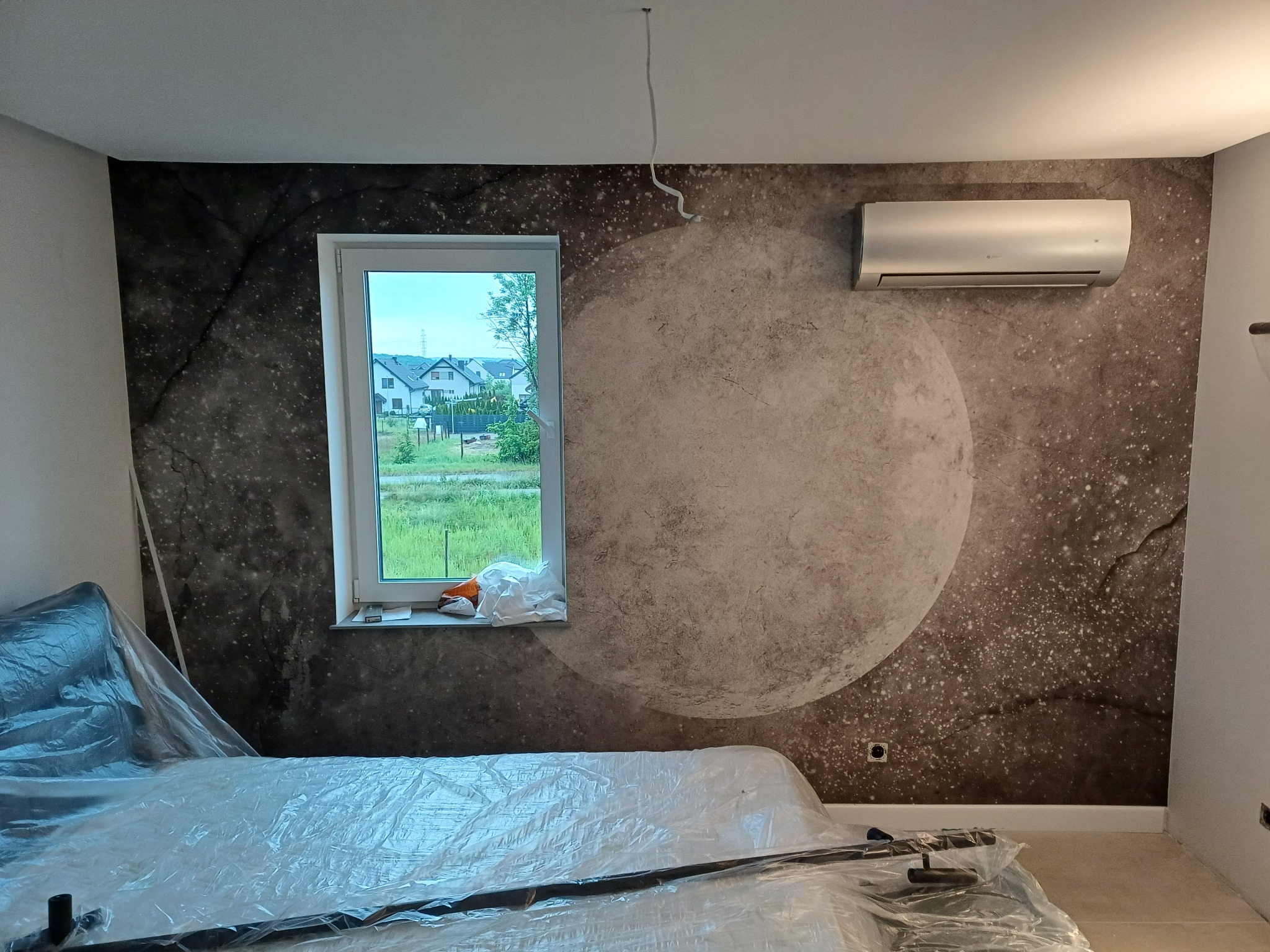 Zamontowany klimatyzator na ścianie z motywem księżyca w pokoju, meble zabezpieczone folią malarską, widok z okna na domy.