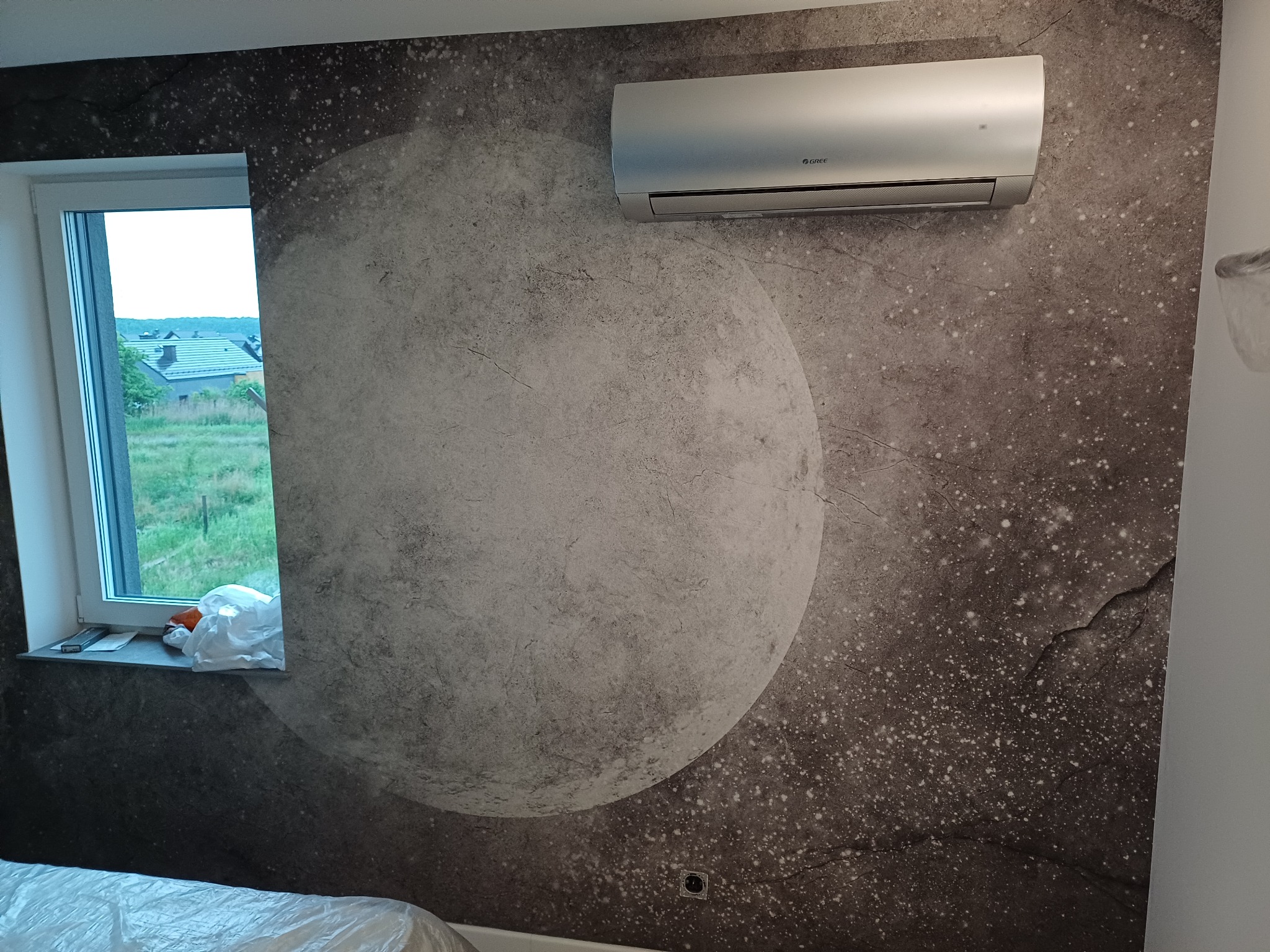 Zamontowany srebrny klimatyzator marki Gree na ścianie z tapetą imitującą księżyc, widok z okna na domy w tle.