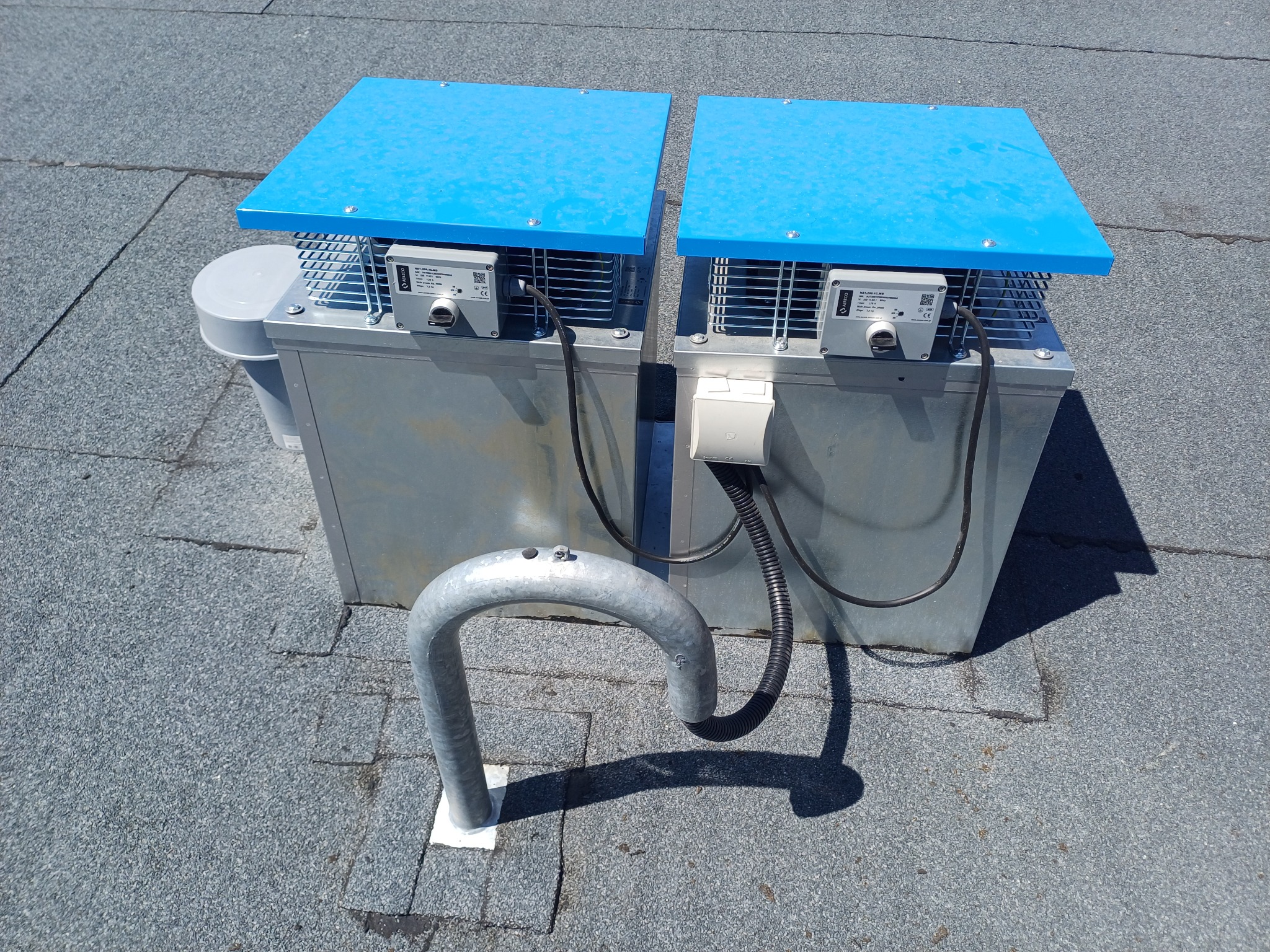 Dwa srebrne, metalowe urządzenia z niebieskimi pokrywami na dachu pokrytym papą, połączone przewodami elektrycznymi i wsparte metalowym pałąkiem.