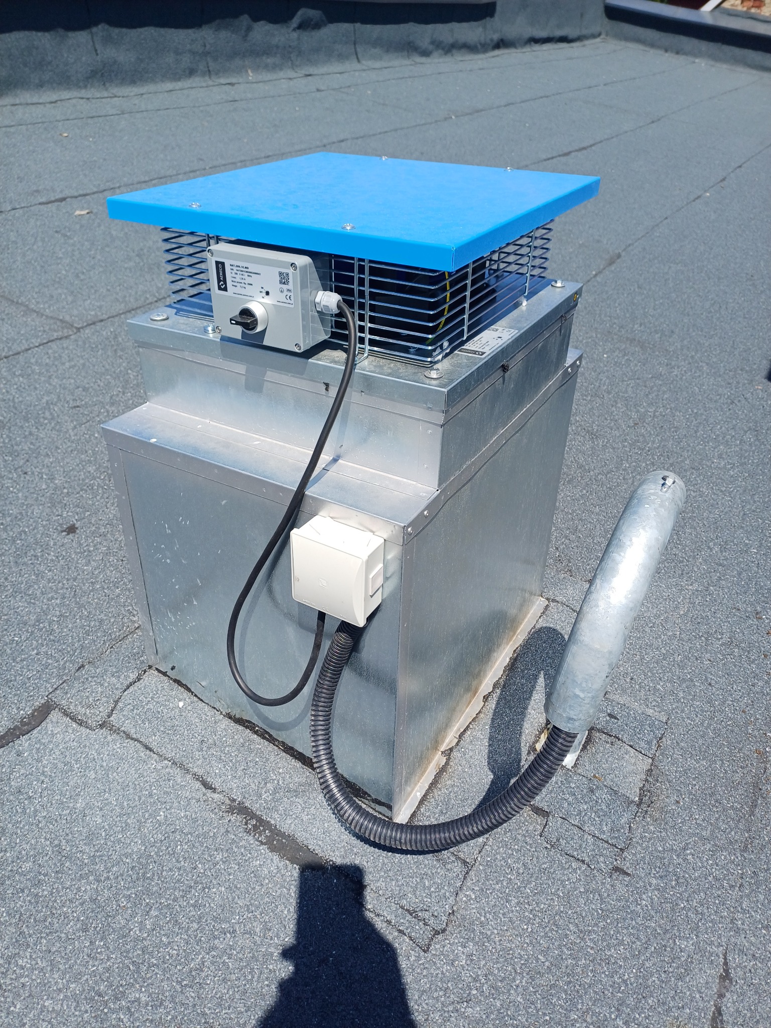 Zewnętrzny moduł rekuperacji z niebieskim daszkiem i metalową obudową na płaskim dachu pokrytym papą.