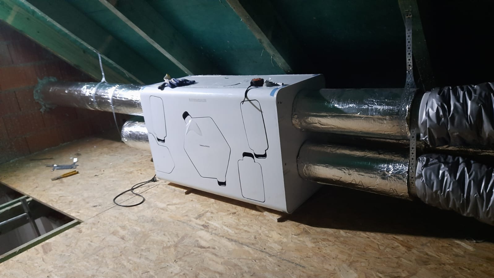 Centrala wentylacyjna z rekuperacją zamontowana na poddaszu, widoczne przewody wentylacyjne w izolacji aluminiowej, narzędzia leżące na podłodze, surowe drewniane wykończenie poddasza.