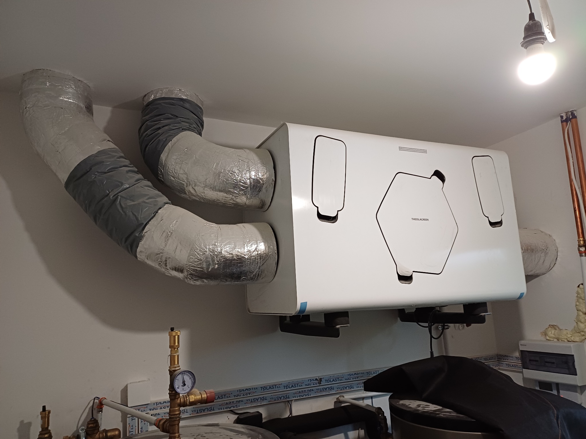 Biała jednostka rekuperacyjna THESSLAGREEN zamontowana pod sufitem, z podłączonymi izolowanymi przewodami wentylacyjnymi, widoczne rury miedziane i manometr.