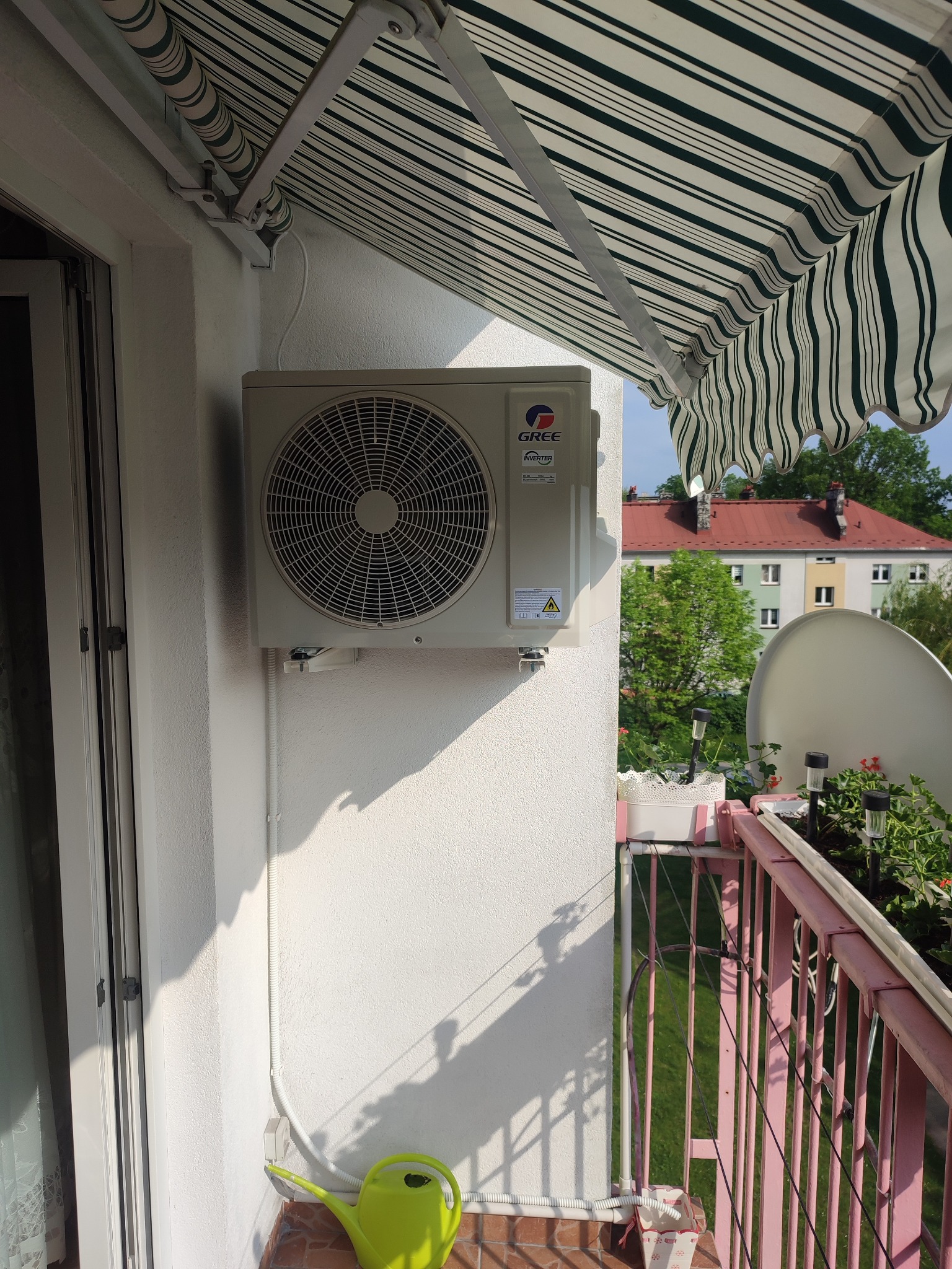 Zewnętrzna jednostka klimatyzacji marki Gree zamontowana na balkonie, widoczne osłony przeciwsłoneczne, kwiaty w donicach i budynki w tle.