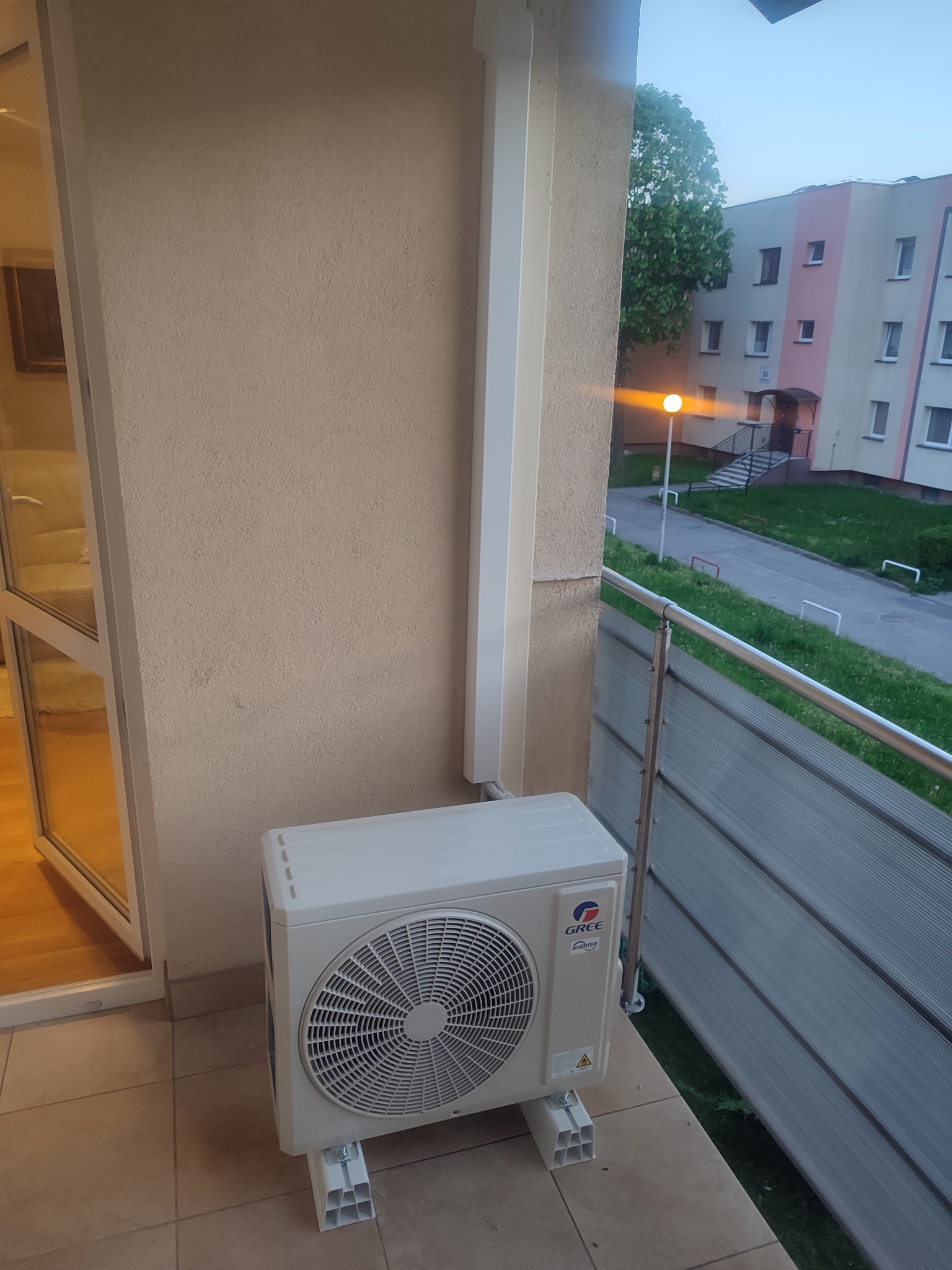 Zewnętrzna jednostka klimatyzacji GREE zamontowana na balkonie mieszkania w bloku, widok na osiedle mieszkaniowe i latarnię uliczną.