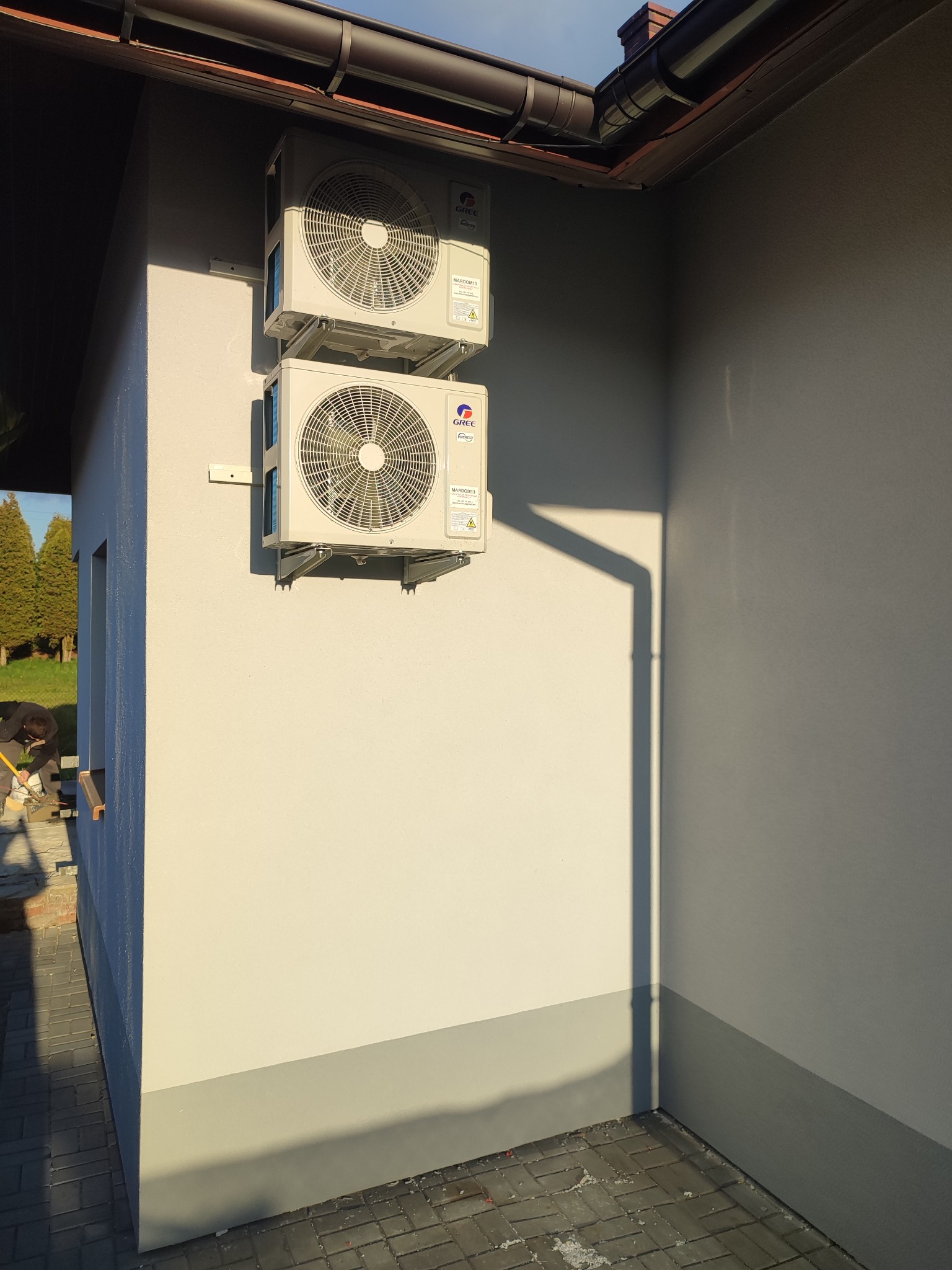 Dwa agregaty klimatyzacyjne marki Gree zamontowane jeden nad drugim na jasnej elewacji budynku, widoczny fragment rynny i cień, w tle zieleń drzew
