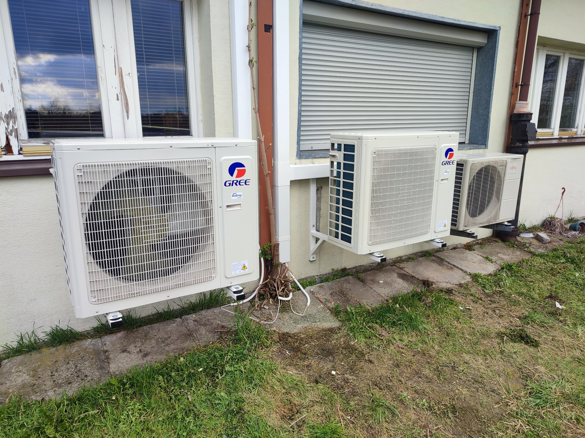 Trzy jednostki zewnętrzne klimatyzacji zamontowane na elewacji budynku z widocznymi przewodami i elementami mocującymi, trawa i kostka brukowa u podstawy.