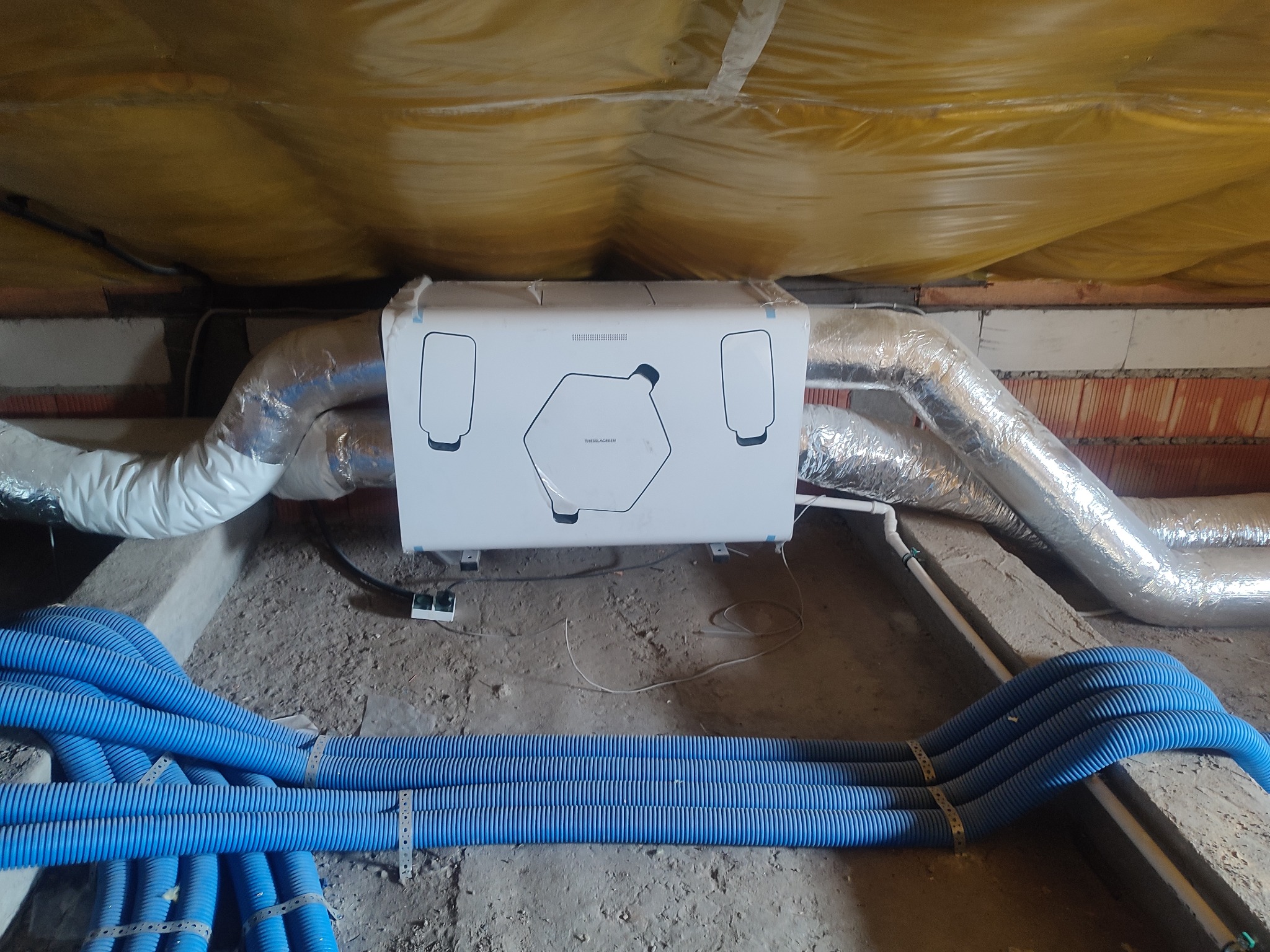 Centrala wentylacyjna z rekuperacją, biała obudowa z czarnymi elementami, podwieszona pod stropem, widoczne niebieskie rury wentylacyjne i srebrne przewody izolowane termicznie, strop ocieplony...