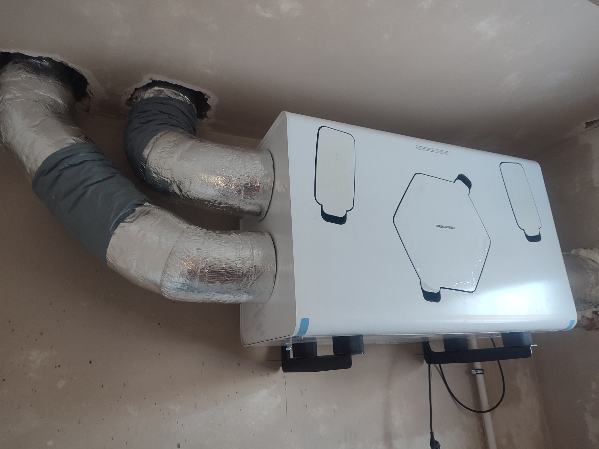 Biała jednostka rekuperacyjna marki Thessla Green zamontowana pod sufitem, z podłączonymi aluminiowymi przewodami wentylacyjnymi w izolacji, widoczne niedokończone wykończenie ścian.