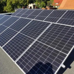 Rząd paneli fotowoltaicznych z białymi ramami zamontowany na płaskim dachu pokrytym papą, widok z góry w słoneczny dzień.