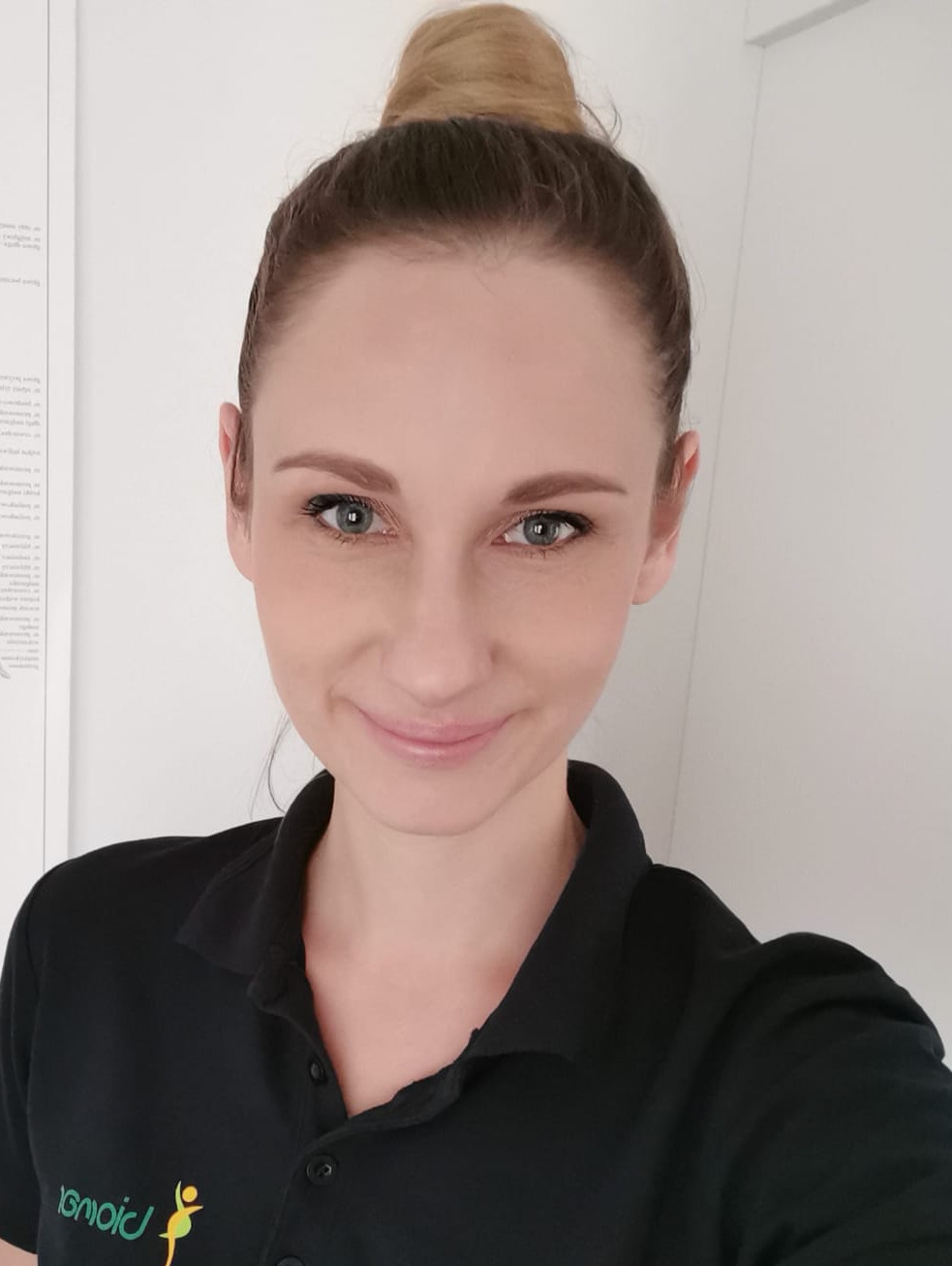 Portret uśmiechniętej kobiety z włosami upiętymi w kok, ubrana w czarną koszulkę polo z logo przedstawiającym sylwetkę osoby w ruchu.