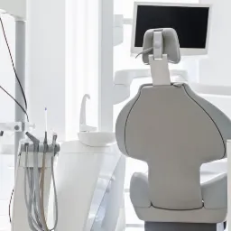 Jasne, sterylne wnętrze gabinetu stomatologicznego z fotelem dentystycznym, lampą zabiegową, monitorem, zestawem narzędzi i modelem uzębienia na białej szafce.