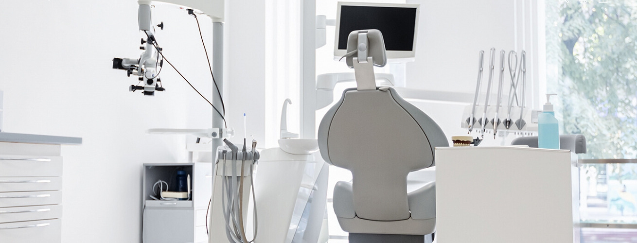 Jasne, sterylne wnętrze gabinetu stomatologicznego z fotelem dentystycznym, lampą zabiegową, monitorem, zestawem narzędzi i modelem uzębienia na białej szafce.