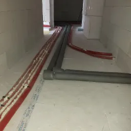 Rury grzewcze czerwone i szare, częściowo zaizolowane pianką montażową, prowadzone wzdłuż ściany z białych bloczków w surowym stanie, widoczne etykiety producenta rur na podłodze.