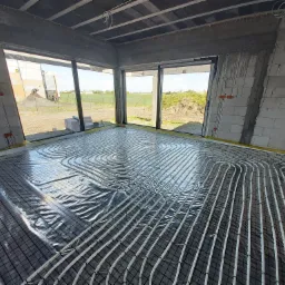 Rozłożone rury ogrzewania podłogowego na czarnej siatce w surowym wnętrzu budynku z widokiem na zewnątrz przez duże okna.