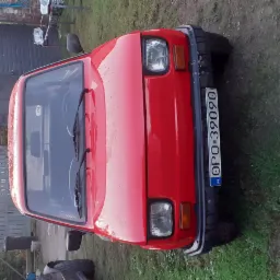 Czerwony Fiat 126p, widok z przodu, z widoczną tablicą rejestracyjną na tle trawnika i budynków gospodarczych.